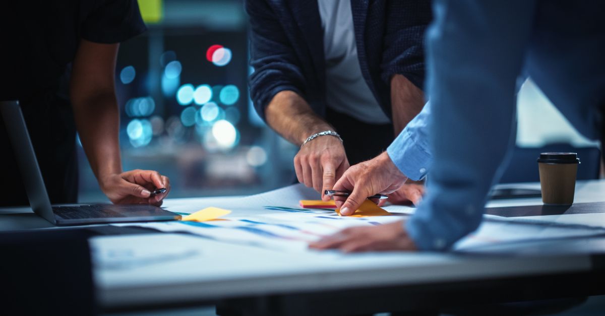 A team of professionals collaborating around a desk, reviewing reports and data on laptops and printouts — representing how organizations address Midmarket ERP challenges by transitioning from legacy systems to modern cloud-based solutions for better efficiency and growth.
